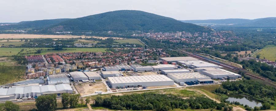 Letecký pohľad na moderný priemyselný park s viacerými skladovými halami. V pozadí sa nachádza obec, železničná trať a zalesnený kopec.