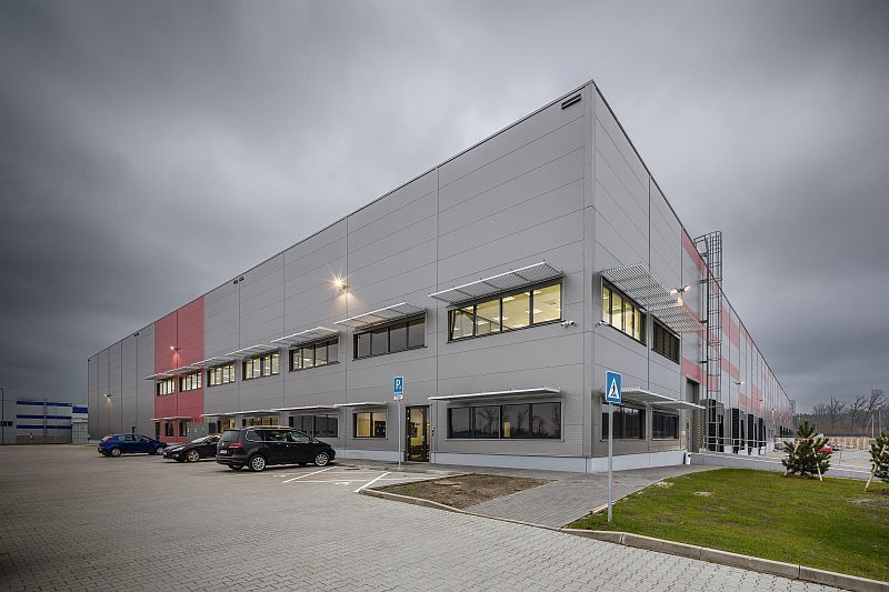 Moderná priemyselná hala zo šedých a červených panelov s administratívnou časťou, parkoviskom a zaparkovanými autami pod zamračenou oblohou.