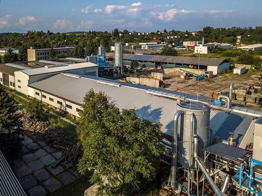 Letecký pohľad na priemyselný areál s výrobnými halami, kovovými silami, potrubnou vzduchotechnikou a spevneným dvorom.