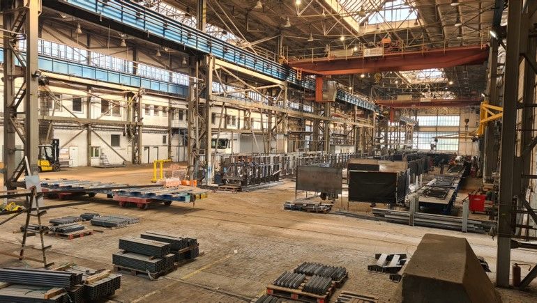 Interiér veľkej priemyselnej výrobnej haly s oceľovou konštrukciou, mostovým žeriavom a uskladneným kovovým materiálom na podlahe.