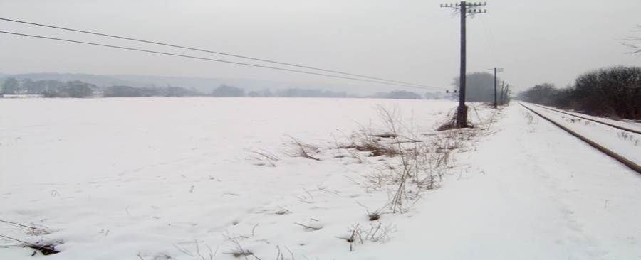 Zasnežený rovinatý pozemok v zime, popri ktorom vedie železničná trať a stĺpy elektrického vedenia.