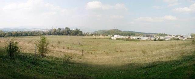 Panoramatický pohľad na rozsiahly trávnatý pozemok s priemyselnými budovami a kopcami v pozadí.
