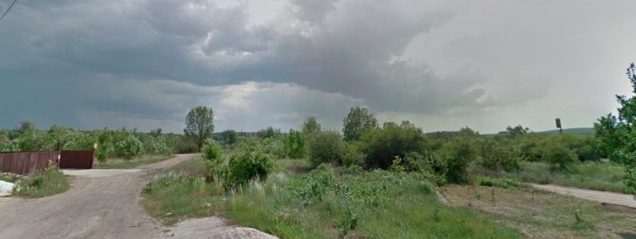Nezastavaný trávnatý pozemok s prašnou prístupovou cestou, oplotením a okolitou zeleňou pod zamračenou oblohou.
