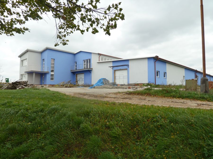 Modro-sivá administratívno-priemyselná budova s prízemnou halou, garážovými bránami a priľahlým trávnatým pozemkom.